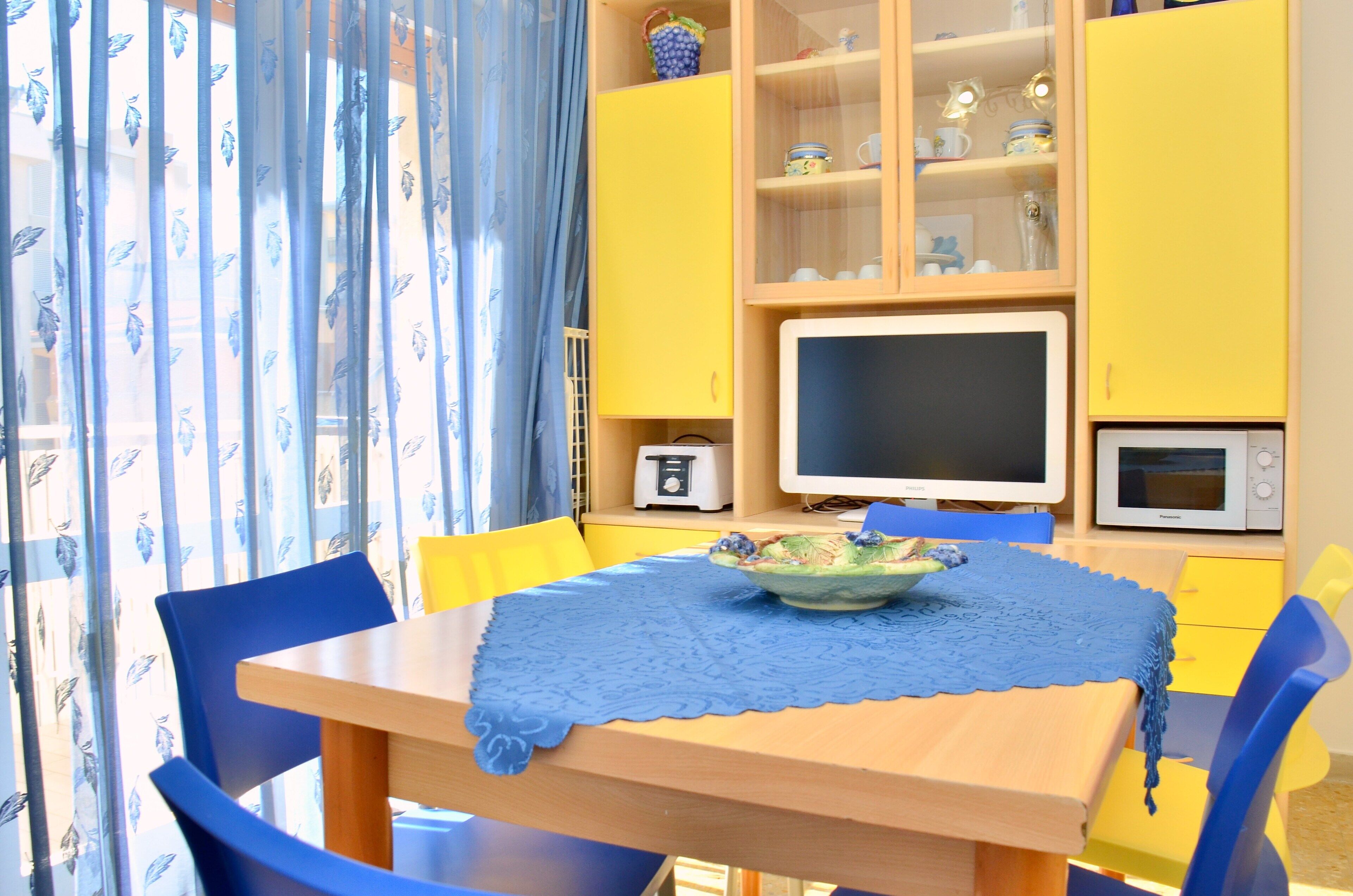 A dining area with a table, chairs, and a blue tablecloth. There are yellow cabinets with glass doors, a microwave, and a toaster.