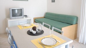 Ein kleiner, ordentlicher Raum mit einem Esstisch für zwei Personen, einem grünen Sofa, einem Fernseher auf einem weißen Schrank und zwei gerahmten Bildern an der Wand.