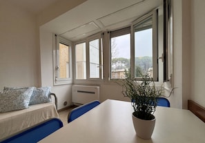 Een kamer met een witte tafel, blauwe stoelen, een kamerplant, een bank met opvallende kussens en grote ramen met jaloezieën.