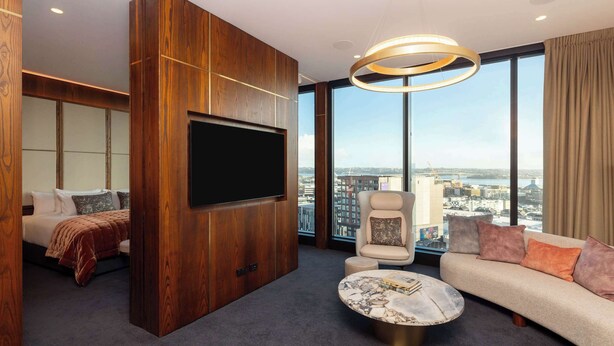 A modern hotel room with a large TV, a sofa, a chair, and a view of the city. - Horizon by SkyCity  (Auckland)