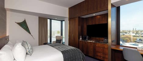 A modern hotel room with a large bed, a desk with a flat-screen TV, and a city view.