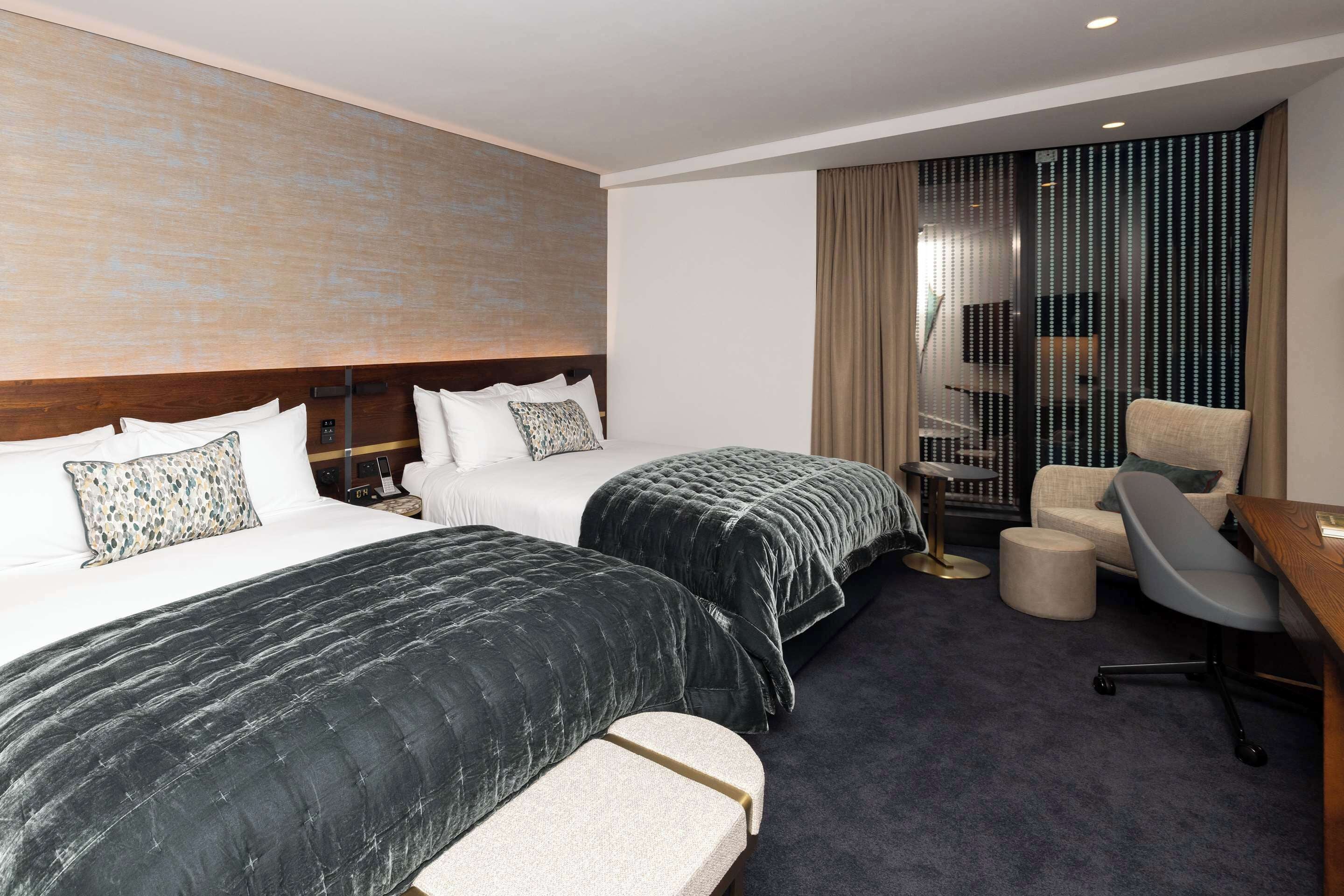 A hotel room with two beds, a desk, a chair, and a large window with curtains.