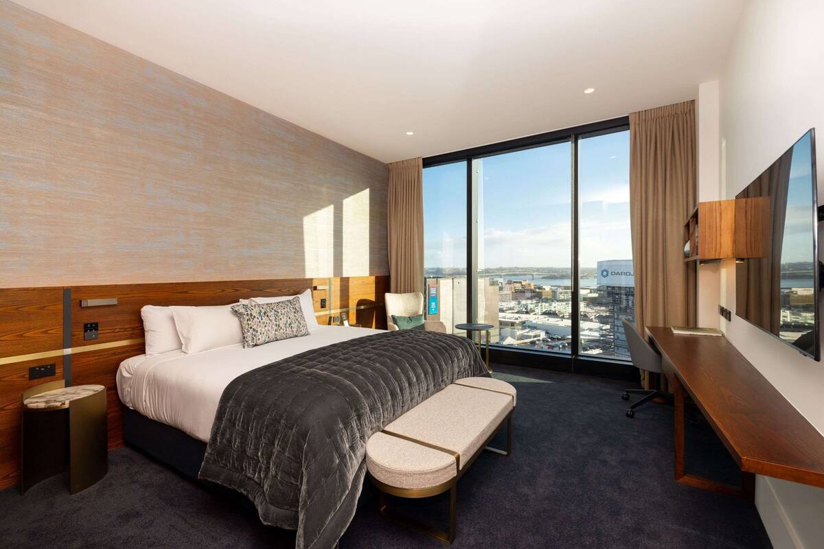 Une chambre d’hôtel moderne avec un grand lit, un bureau, un banc et une vue sur la ville.