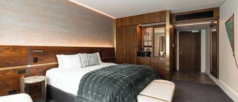 Ein modernes Hotelzimmer mit einem großen Bett, einem Nachttisch, einem kleinen runden Tisch, einer Bank und einem Kleiderschrank.