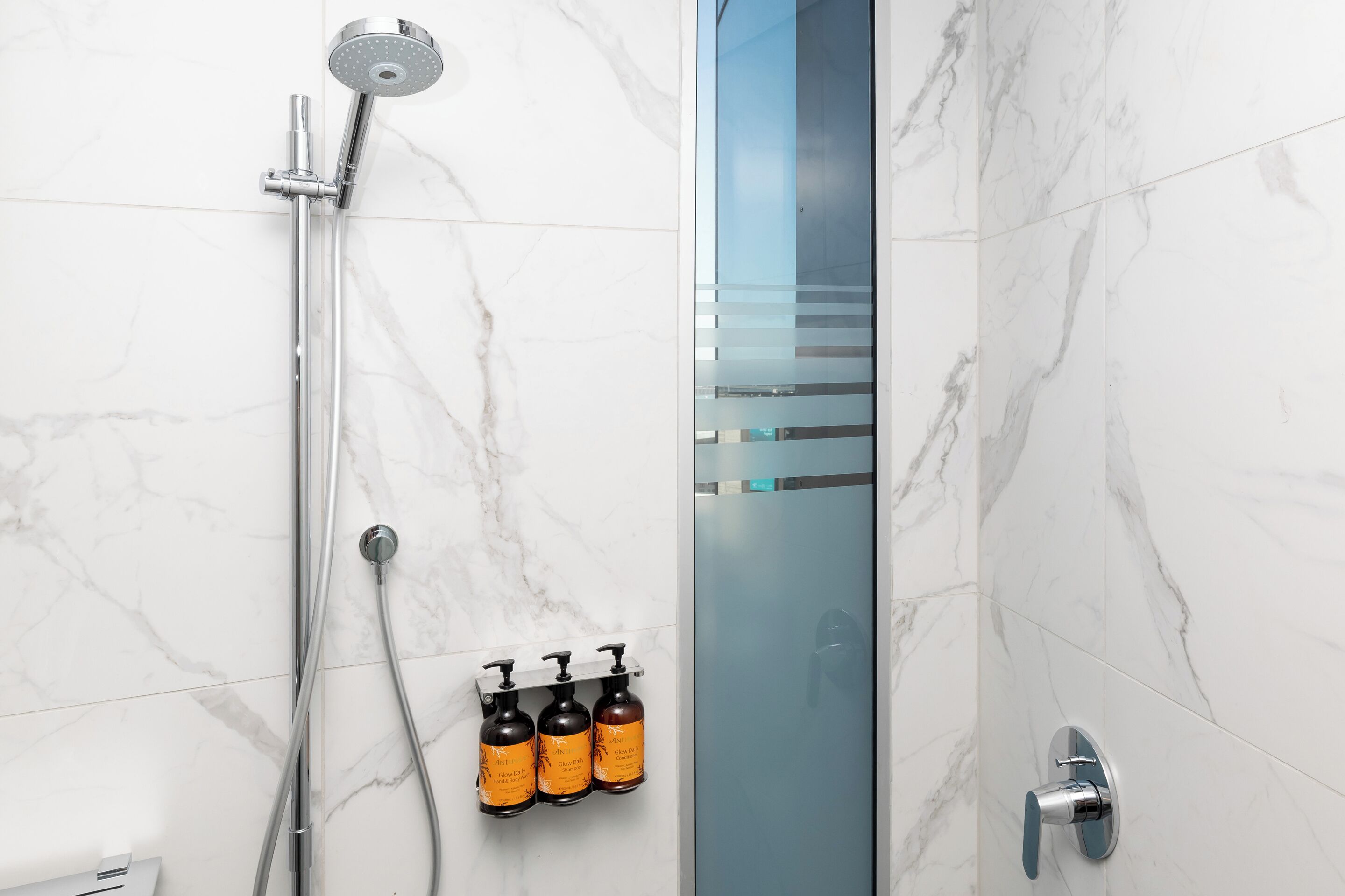 Un baño moderno con cabina de ducha de vidrio, ducha de mano y tres botellas de champú y acondicionador fijadas a la pared.