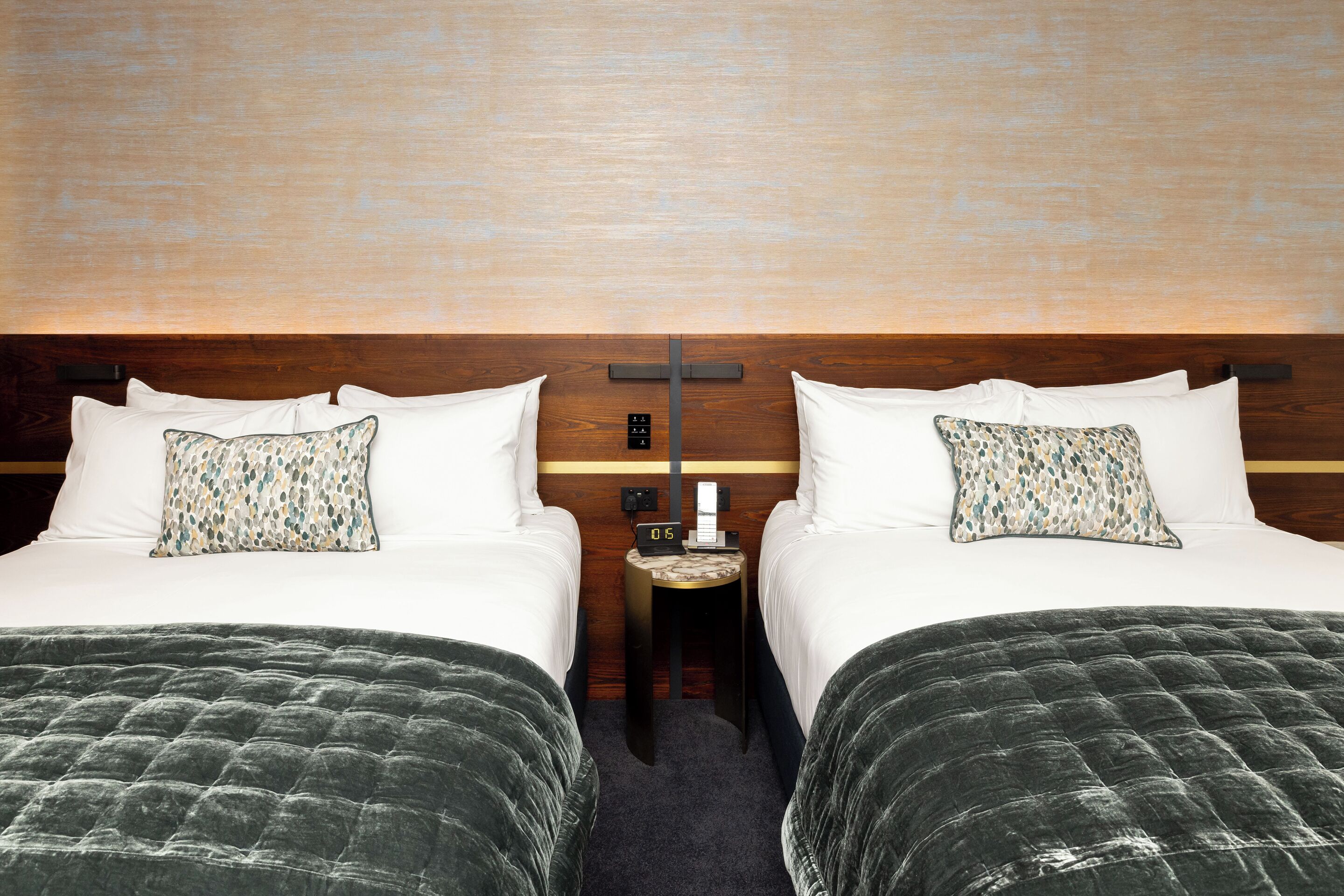 Quarto de hotel com duas camas, uma mesa de cabeceira e um relógio na parede.