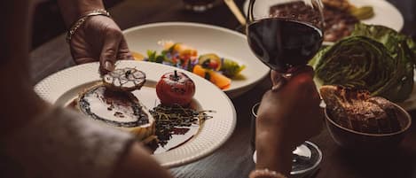 A dining table with plates of food, a bowl of bread, and glasses of red wine.