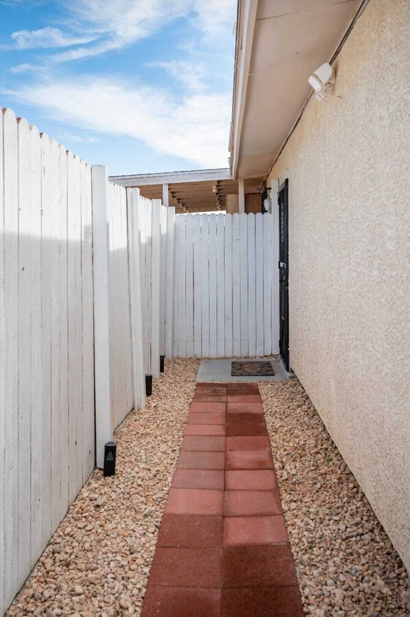 A narrow pathway with a red brick border leading to a door, flanked by white wooden fences and a beige wall. - Private Studio in Las Vegas with Two Beds, Kitchenette & Parking (Las Vegas)
