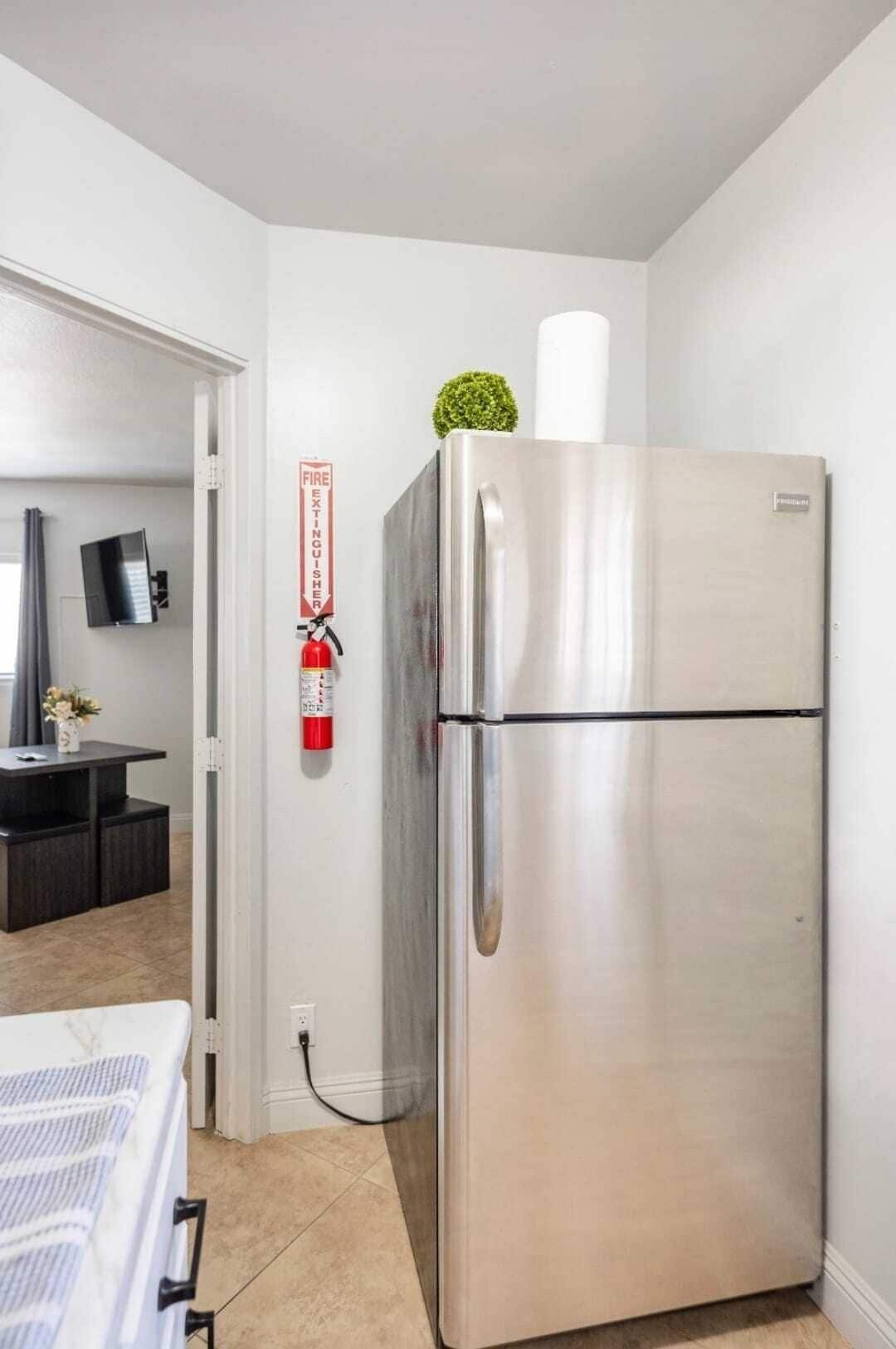 A compact kitchen with a stainless steel refrigerator, a small potted plant on top, and a fire extinguisher mounted on the wall.