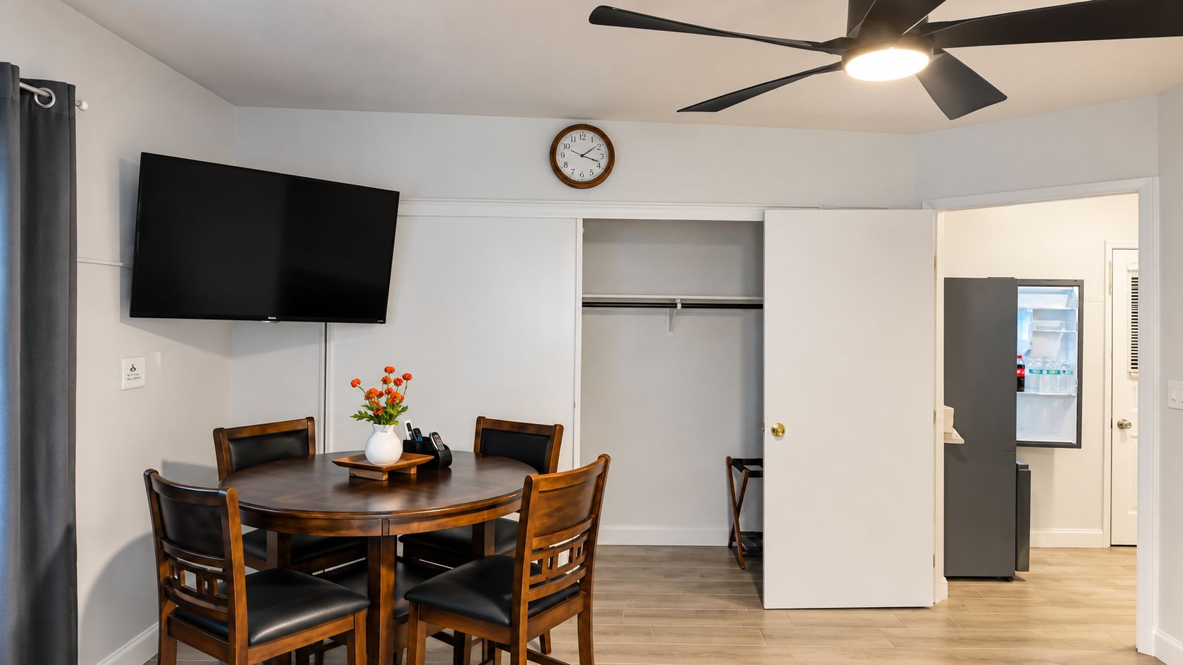 A dining area with a round table and chairs, a clock on the wall, a ceiling fan, and a door leading to a kitchen.