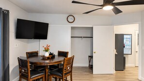A dining area with a round table and chairs, a clock on the wall, a ceiling fan, and a door leading to a kitchen.