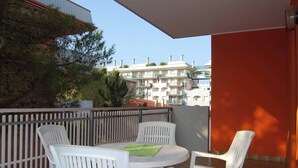 A balcony with white plastic chairs and a table, a wooden fence, and a view of buildings in the background. - Warm Two-room Flat With Balcony Near the Beach (San Michele al Tagliamento)