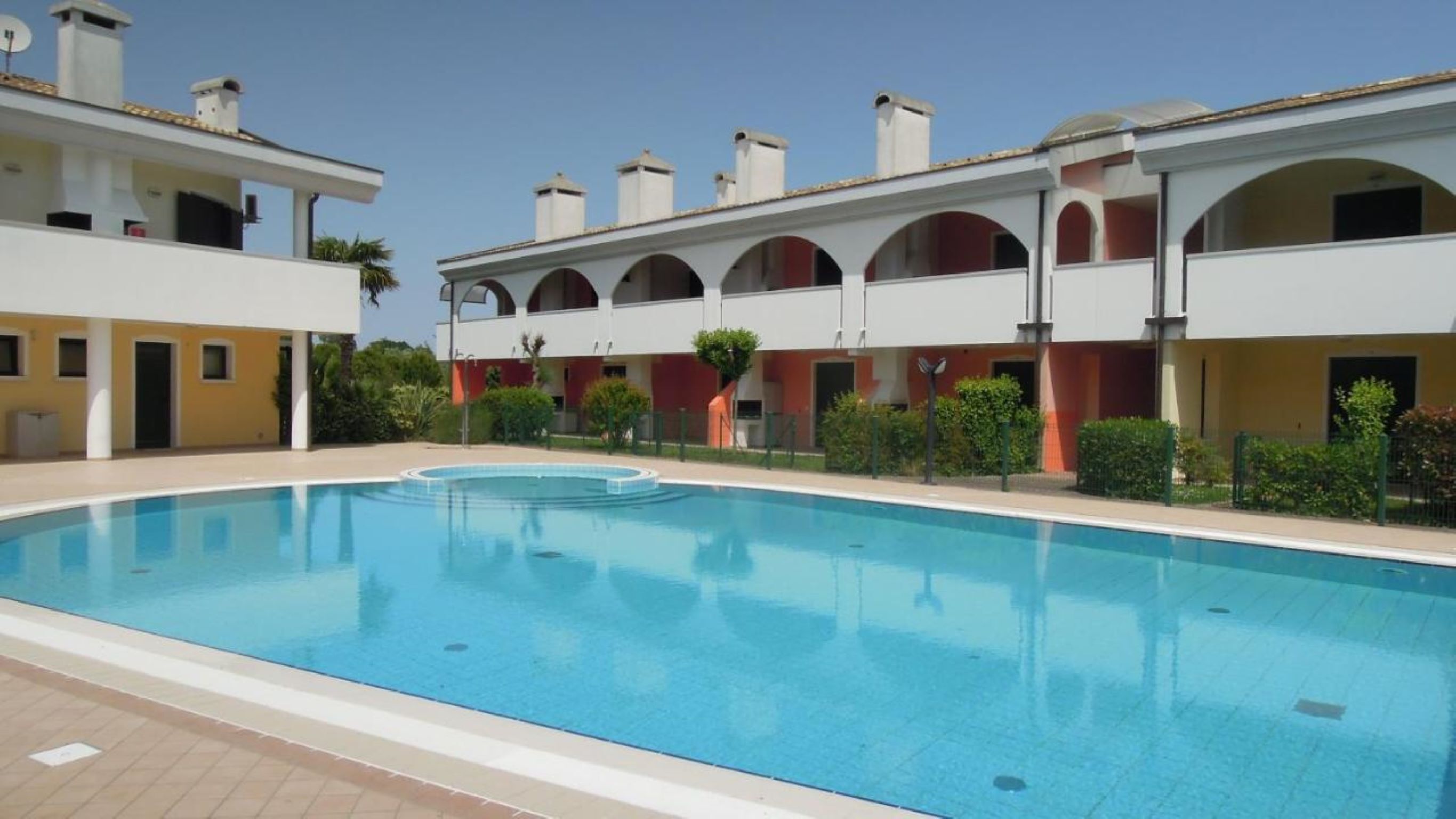 A swimming pool with a tiled deck, a covered outdoor area with multiple arches, and a building with balconies and chimneys.