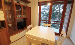 A dining area with a white table and chairs, a wooden cabinet with glass doors, and a view through a glass door to the outside. - Spacious Apartment in Lignano Pineta (Lignano Sabbiadoro)