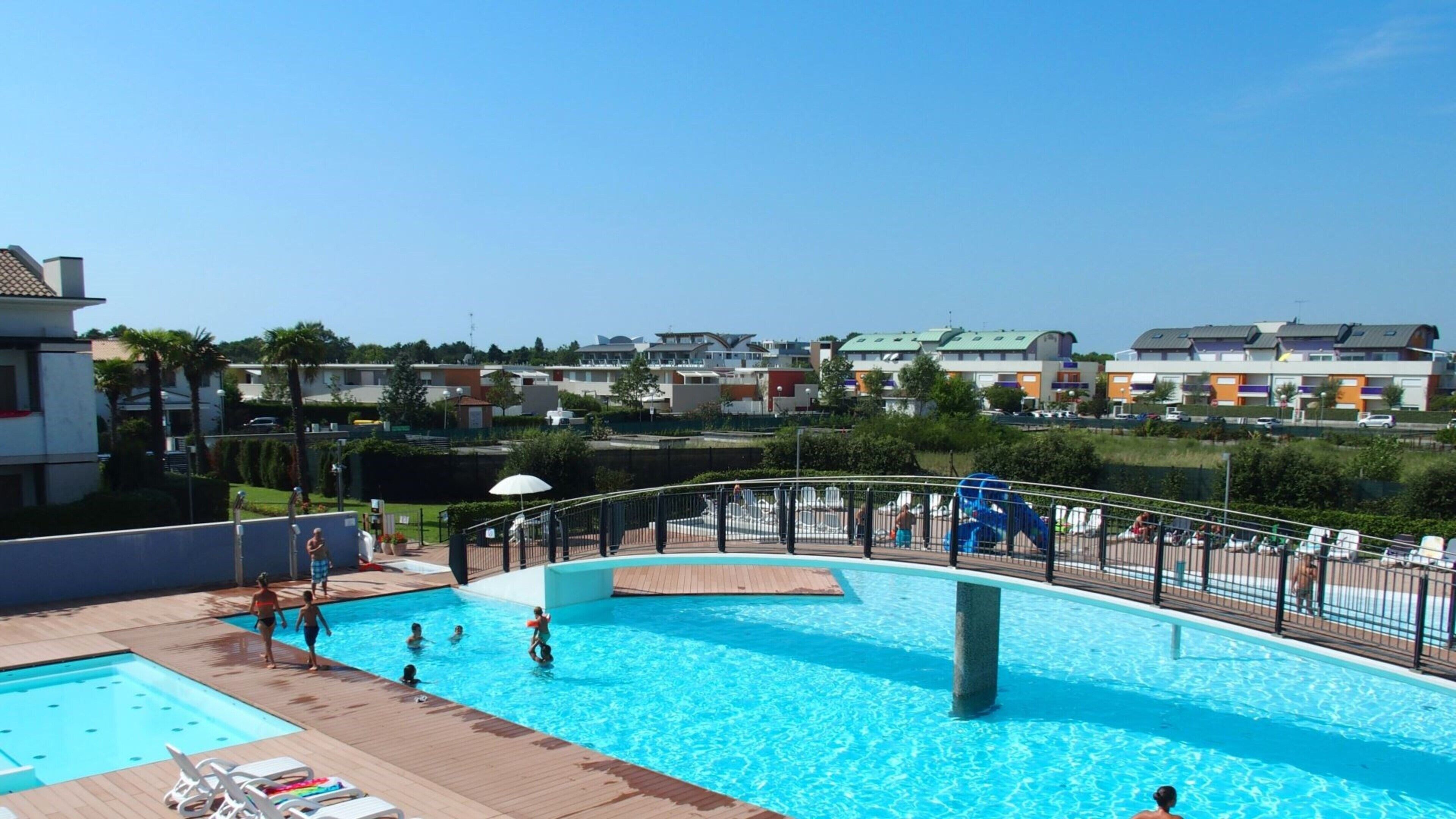 A swimming pool with a wooden deck, surrounded by a metal fence, and overlooking a residential area with various buildings.
