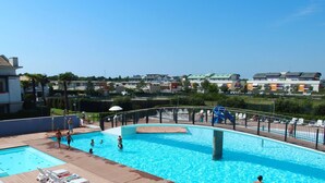 Ein Schwimmbecken mit Holzterrasse, umgeben von einem Metallzaun, mit Blick auf eine Wohngegend und verschiedenen Gebäuden.