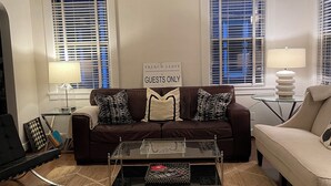 A living room with a brown sofa, a glass coffee table, and a beige couch. There are two windows with blinds, a ceiling fan, and a sign that reads "French Leave Guests Only". - Masters rental - Designer MCM updated bungalow   (Augusta)
