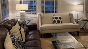 A living room with a brown leather sofa, a beige sofa with a black and white patterned pillow, and a glass coffee table. There are two windows with blinds, a lamp on a side table, and a door in the background. - Masters rental - Designer MCM updated bungalow   (Augusta)