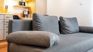 En grå sofa med pude, et køkkenområde med skabe og kaffemaskine, samt en hvid væg med lyskontakt.