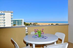 A balcony with a table and chairs, overlooking a beach and a building. - Beautiful Studio Apartment in a Seafront Building (San Michele al Tagliamento)