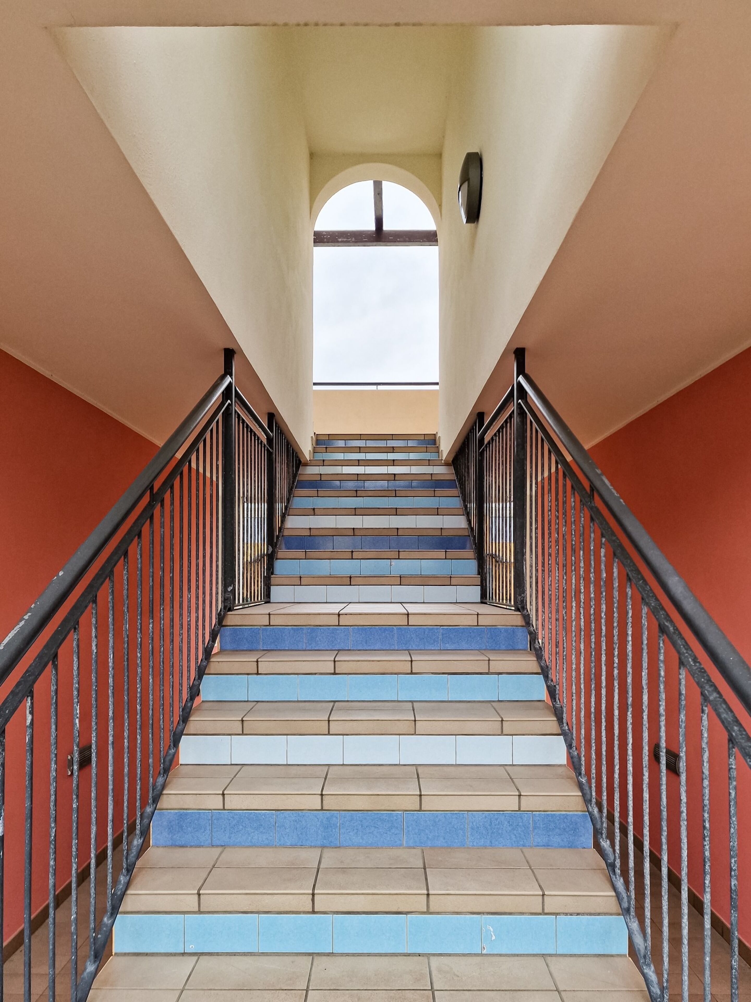 Una escalera con peldaños alternados en azul y beige, barandillas negras y una vista del cielo a través de un ventanal arqueado en la parte superior.