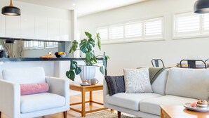 Un salon moderne avec un canapé, un fauteuil et une table basse. On y trouve également un coin cuisine avec des placards et une plante.