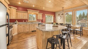 A spacious kitchen with wooden cabinets, a large island with bar stools, and a view of trees through the windows. - Elk Ridge Retreat-25m to Yellowstone+hot tub+home theater (island park)