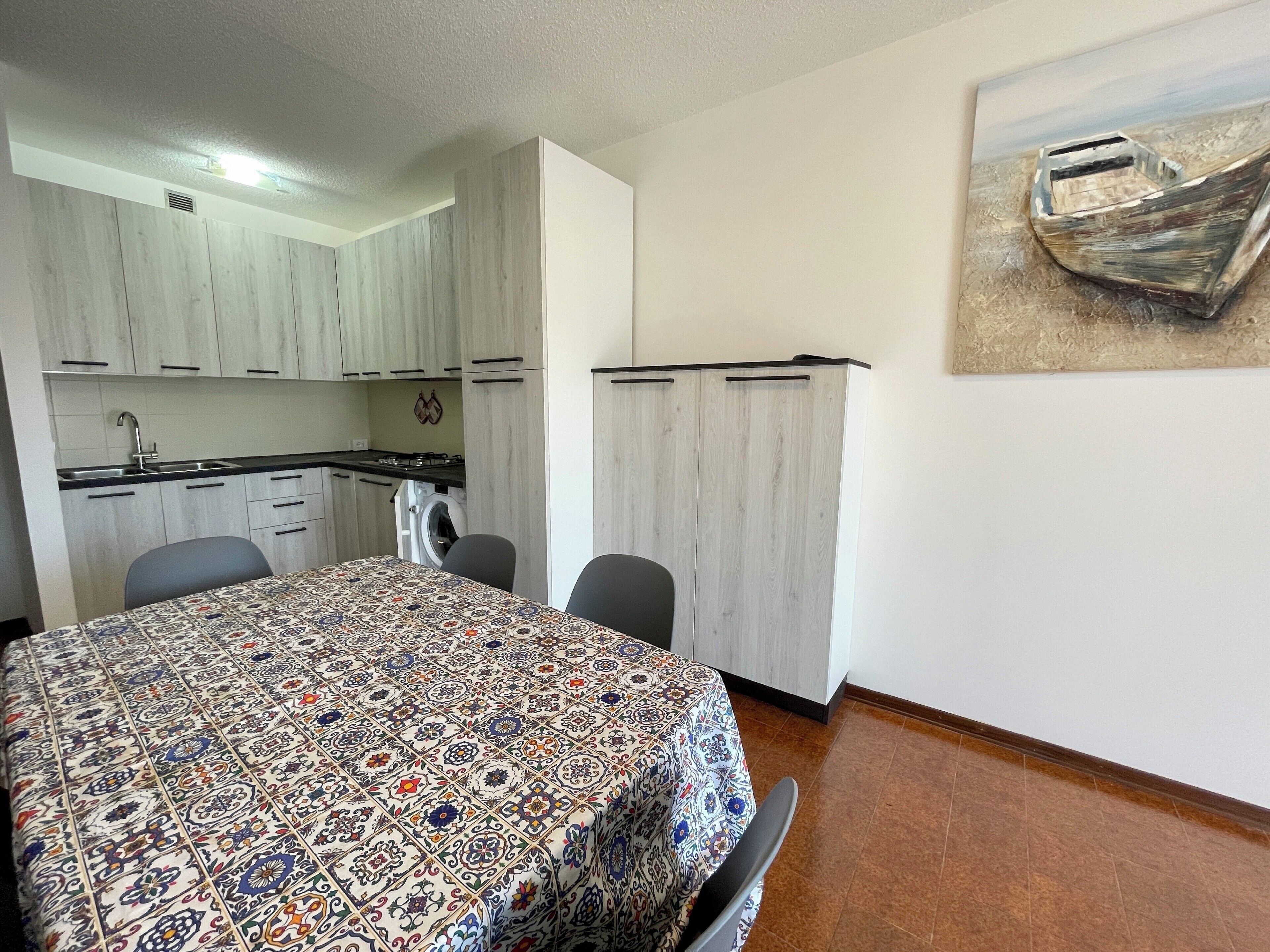 A compact kitchen with white cabinets, a dining table with a patterned tablecloth, and a single chair.