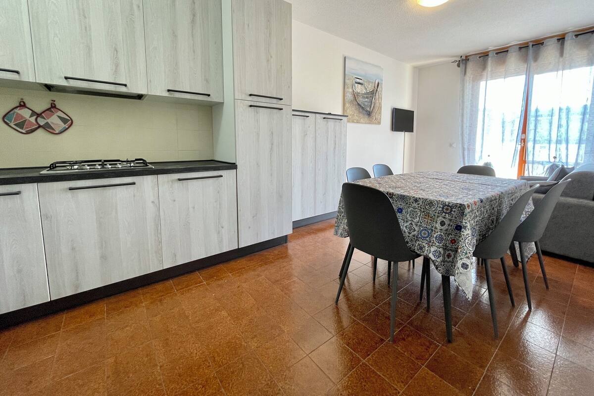 A compact kitchen with white cabinets and a tiled floor, a dining area with a patterned tablecloth and chairs, and a window with curtains.