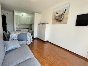 A compact living space with a sofa, a dining table, chairs, a kitchen area, and a mounted television. - Apartment for six With Shared Pool (Caorle)