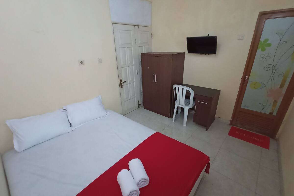 A neatly made bed with white bedding and a red blanket, a wooden wardrobe, a white plastic chair, a small desk, and a television on the wall.