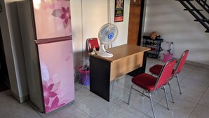 Una cocina pequeña con un refrigerador rosa, una mesa de madera, dos sillas rojas, un ventilador de pie y una escalera al fondo.