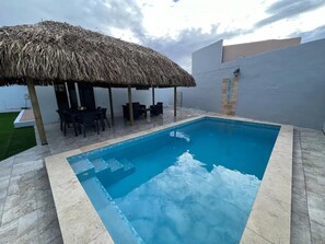 Uma piscina retangular com borda em mosaico, uma estrutura de telhado de palha e área de estar externa.