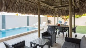 Un espace piscine extérieur doté d’un toit de chaume, de chaises longues et d’une table à manger.