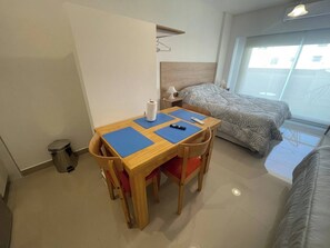 Habitació compacta amb un escriptori de fusta, dos estores blaus, un llit amb roba de llit estampada, una petita taula de nit amb una làmpada i una finestra amb vistes.