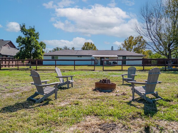 A grassy outdoor area with Adirondack chairs, a fire pit, and a building in the background.