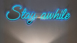 Un letrero de neón que dice 'Stay awhile' sobre una lámpara en una mesita de noche.