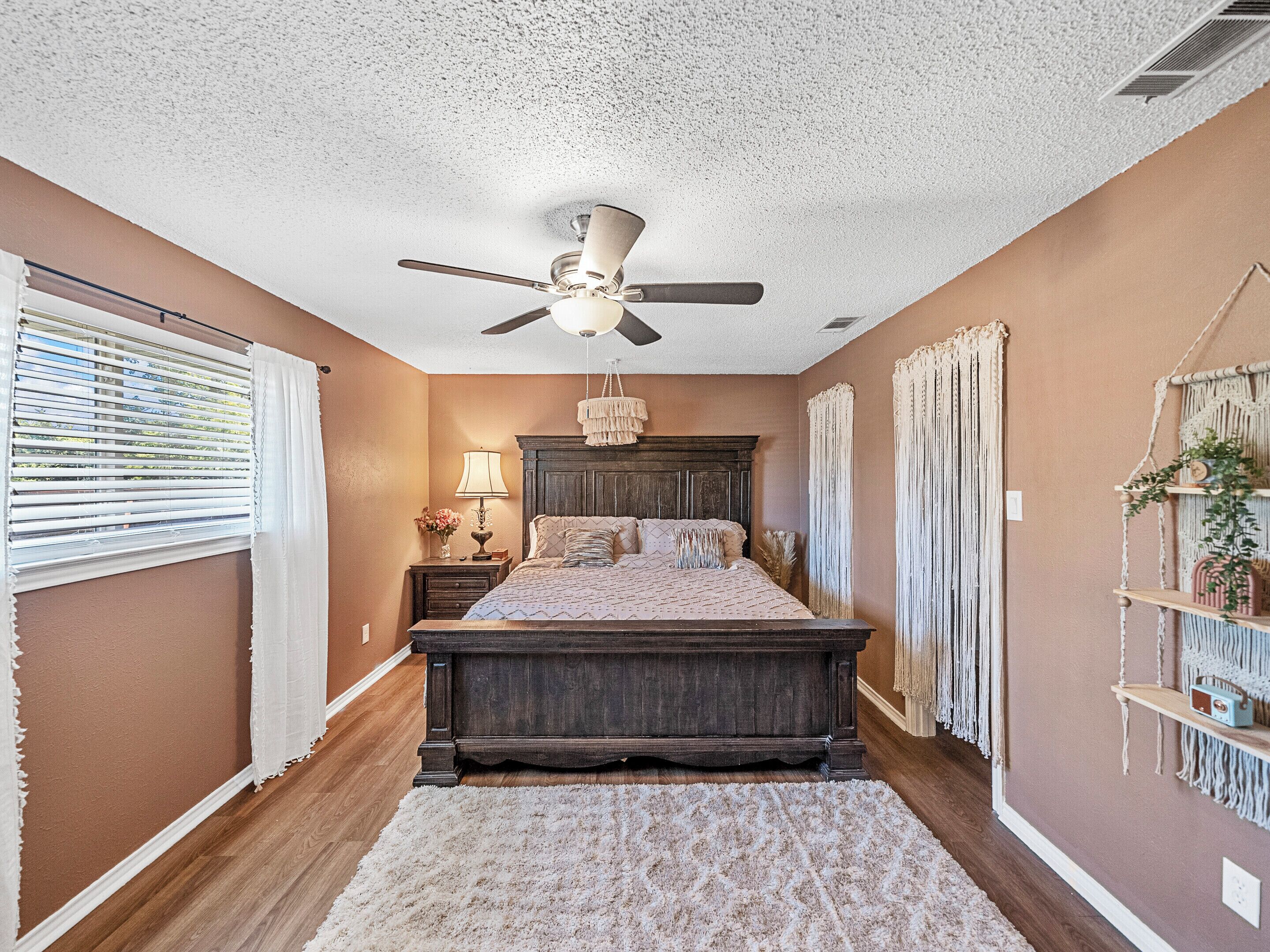 A bedroom with a large bed, a ceiling fan, a nightstand with a lamp, and a macrame wall hanging.
