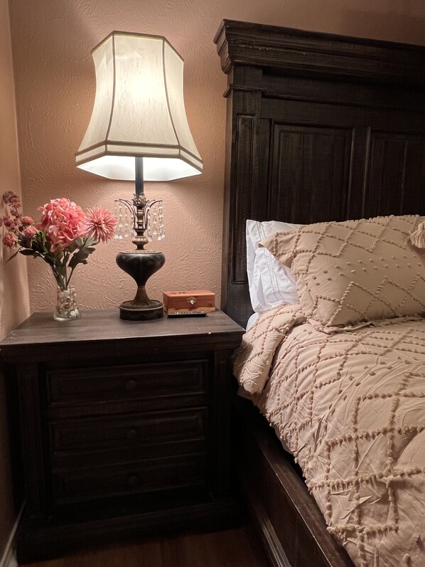 Una cama bien hecha con cubrecama acolchado, una lámpara de noche, un florero con flores y un pequeño cofre de madera sobre una mesita de noche de madera.