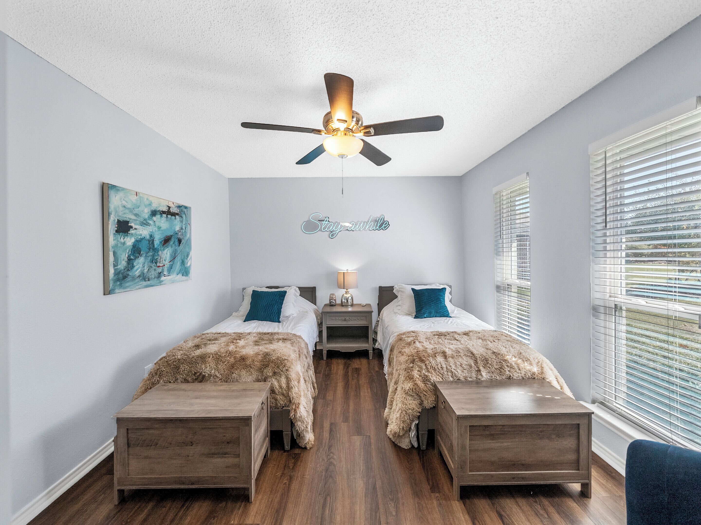 Une chambre avec deux lits, un ventilateur de plafond, un tableau, un panneau et une fenêtre avec des stores.
