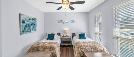 Une chambre avec deux lits, un ventilateur de plafond, un tableau, un panneau et une fenêtre avec des stores.