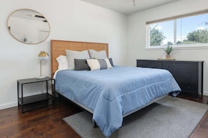 A bedroom with a bed, a nightstand, a dresser, a mirror, and a window with a view of trees.