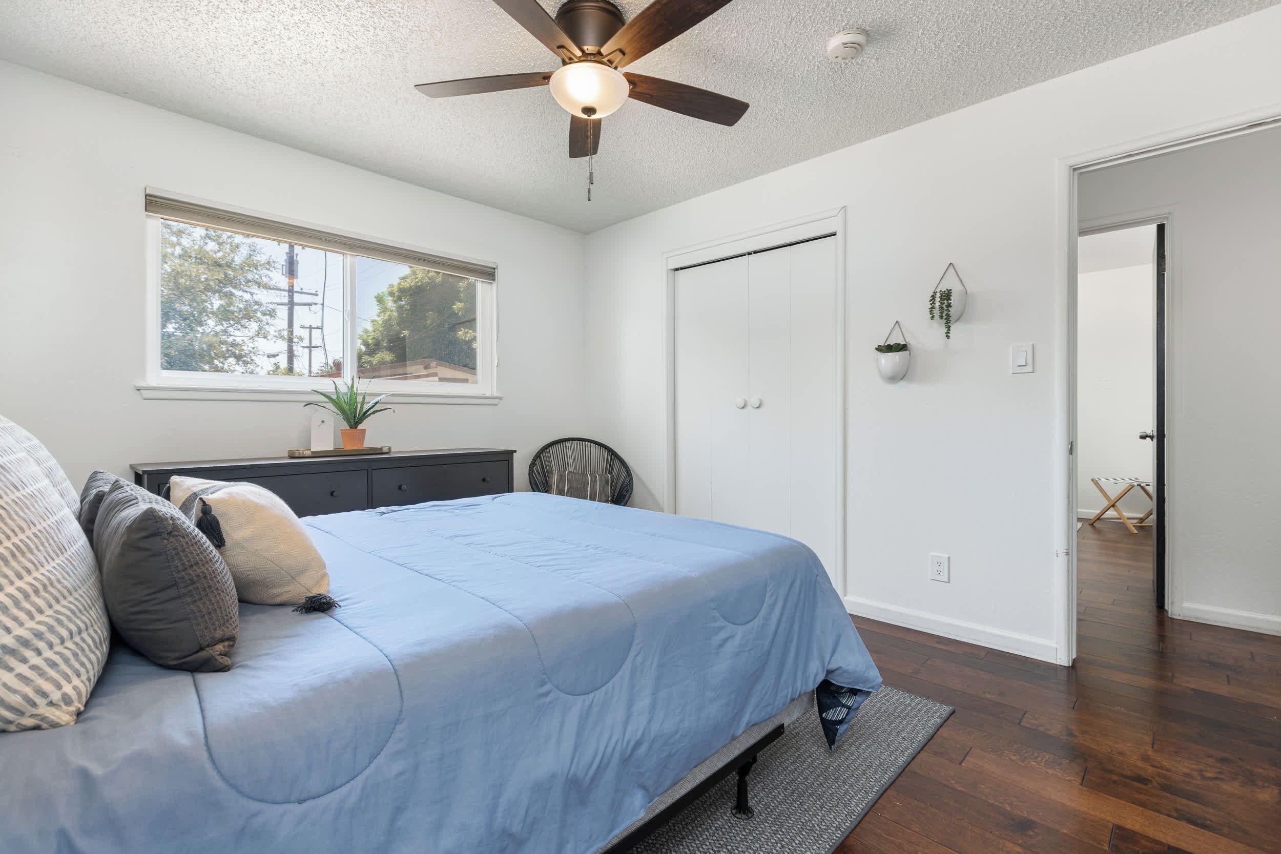 Un dormitorio con cama, ventilador de techo, ventana, puerta y una pequeña planta en maceta.