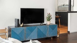 Ein modernes Wohnzimmer mit einem Flachbildfernseher auf einer blau gemusterten Kommode, einer kleinen Pflanze und einem Holzstuhl.