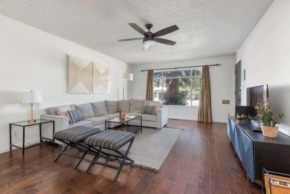 A modern living room with a sofa set, a coffee table, a striped ottoman, a ceiling fan, a TV, and a potted plant.