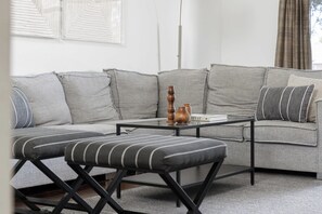 Un salon moderne avec un canapé d’angle gris, un pouf rayé noir et blanc et une table basse en verre agrémentée de vases décoratifs.