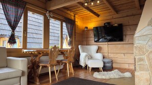 Interior acogedor de una cabaña con una mesa de comedor de madera, sillas, un sillón blanco, un televisor montado en una pared de piedra y una chimenea.