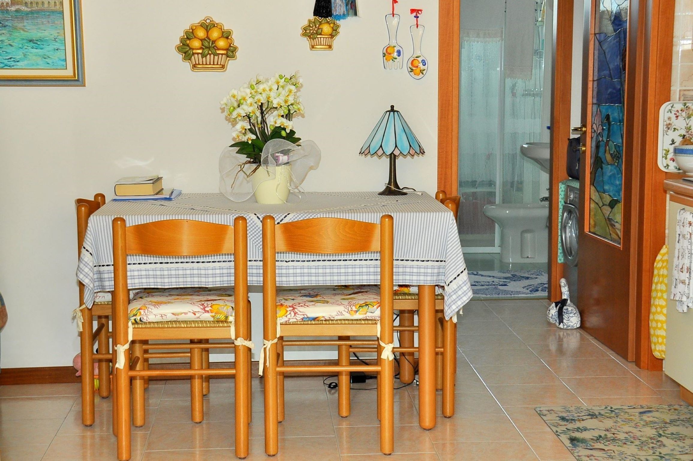 Un comedor con una mesa dispuesta para cuatro personas, rodeada de sillas de madera. En la mesa hay un florero con flores, una lámpara y un cuadro en la pared.