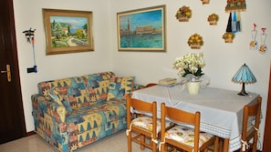 Ein gemütliches Wohnzimmer mit einem farbenfrohen Sofa, einem Esstisch mit Stühlen, einem Blumenstrauß und Wanddekorationen, darunter gerahmte Gemälde und Obstkörbe.