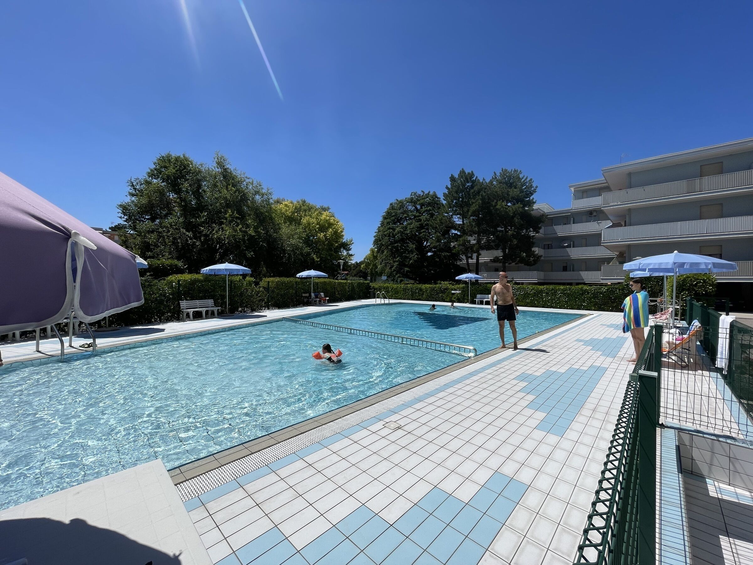 A swimming pool with a tiled deck, surrounded by umbrellas and trees.